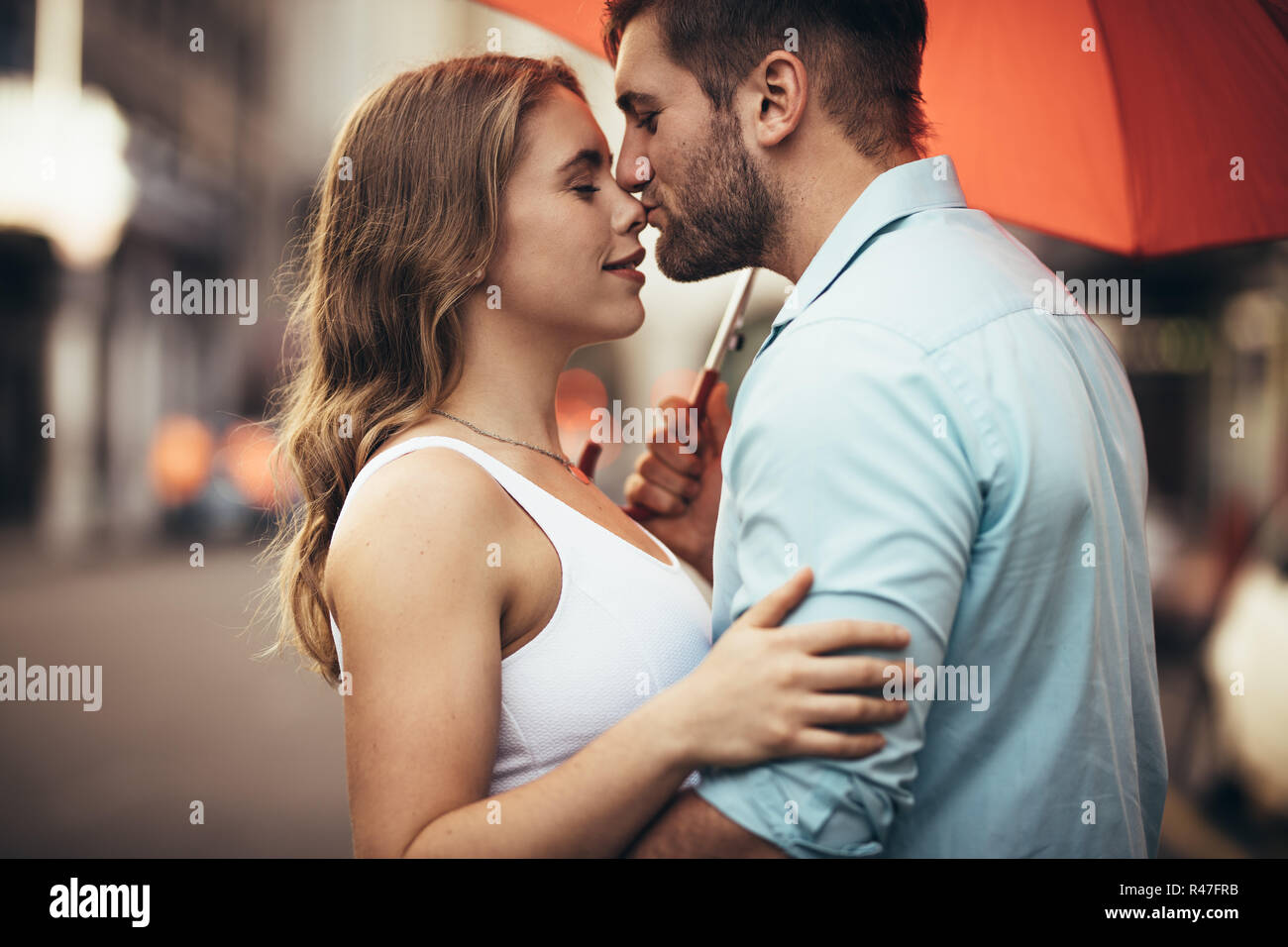 Romantic woman holding les uns les autres avec un parapluie sur rue. Jeune homme embrassant le nez de sa petite amie standing in street embra Banque D'Images