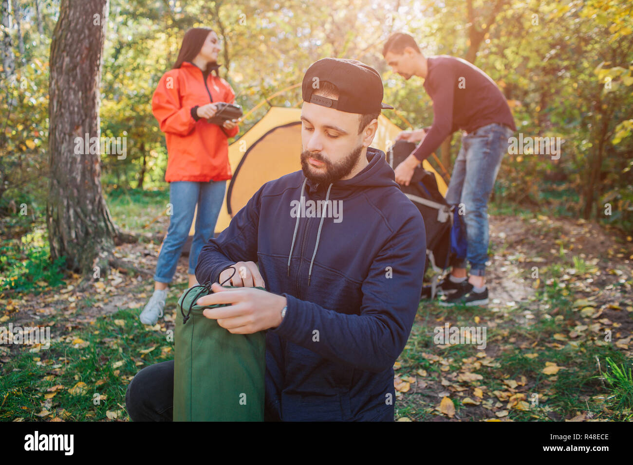 Jeune homme barbu est titulaire d'un sac de couchage et regarde. Il est sérieux. Femme détient la radio dans les mains. Un autre gars travaille avec tente. Banque D'Images