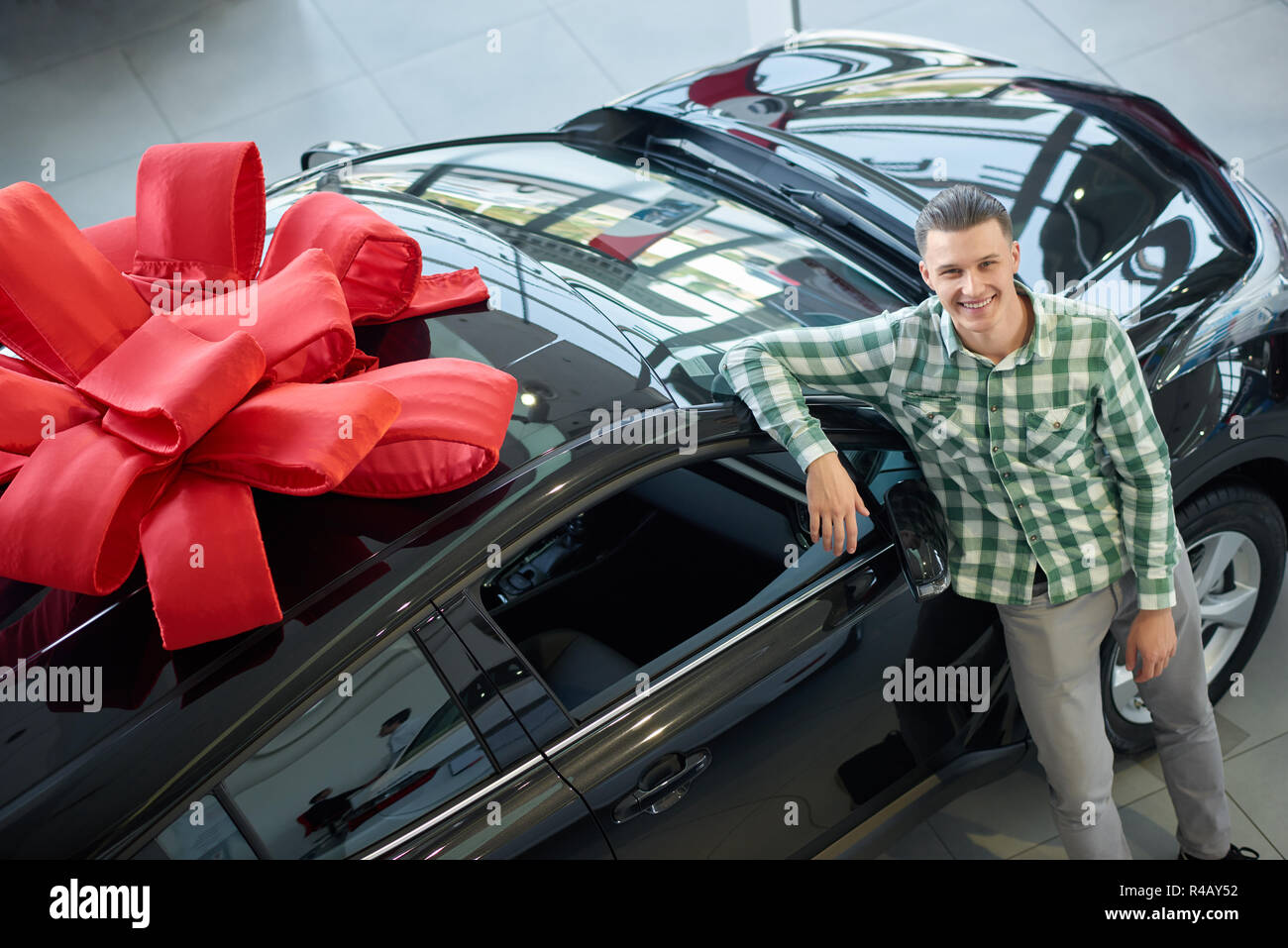 Young man looking at camera et tenir la main sur la prise de voiture ci-dessus. Guy attrayant dans les tenues debout près de Big Black auto avec noeud sur le haut. Heureux gagnant s'appuyant sur son prix, et souriant. Banque D'Images