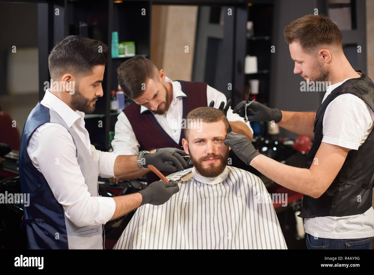 Les jeunes clients masculins sitting in chair et looking at camera tandis que les professionnels de la coiffure et de l'entretien permanent autour de lui à vintage barber shop. Coiffeurs qualifiés coiffer les cheveux et la barbe. Banque D'Images