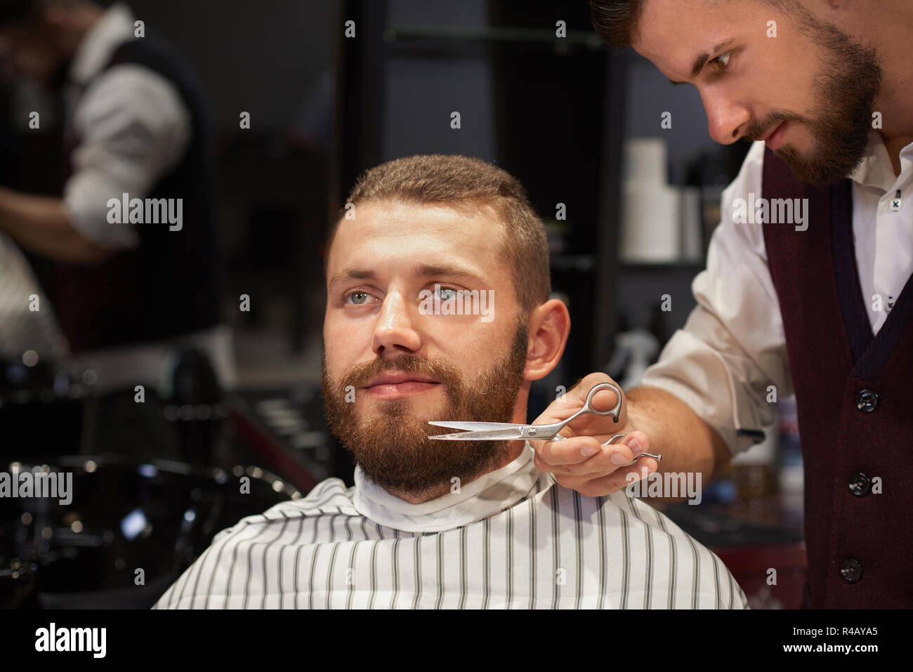 Smiling man sitting in chair dans barber shop, à l'écart et se coupe de barbe. Coiffure qualifiés en chemise blanche et gilet, debout près de son style et de clients masculins avec des ciseaux. Banque D'Images