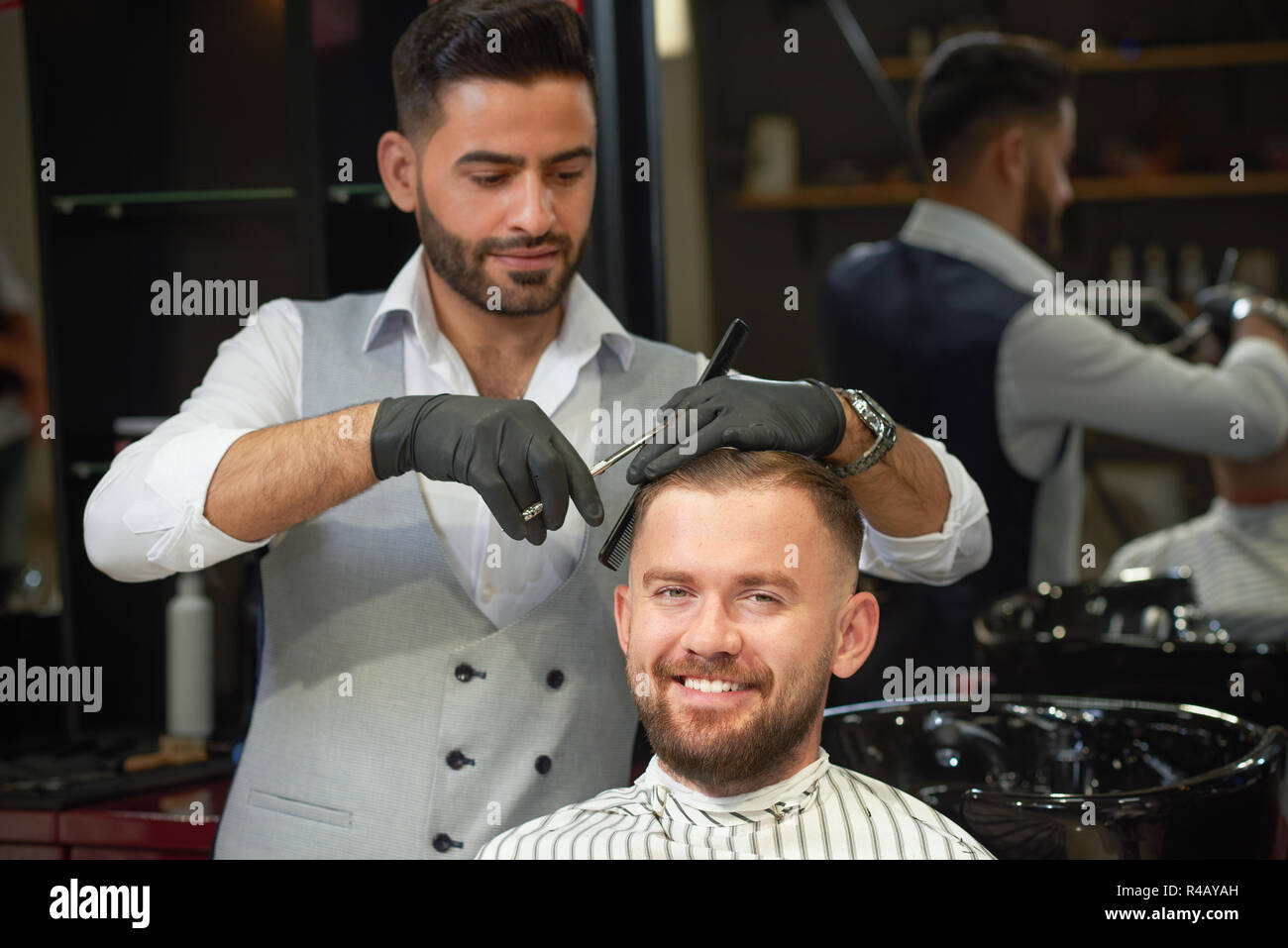 Vue avant de sourire barbu assis dans la chaise dans un salon de coiffure et de mise en plis. Coiffure qualifiés en chemise blanche et gilet coupe élégante faire avec des ciseaux et peigne. Banque D'Images