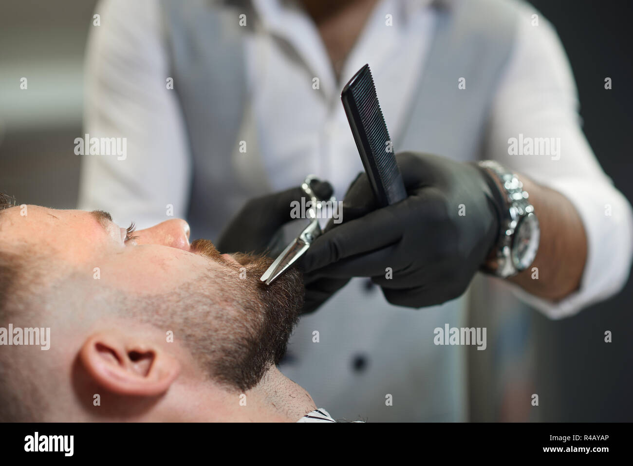 Gros plan du processus de barbe coupe de clients masculins dans barber shop. Coiffure qualifiés en noir gants de protection conforme ciseaux et peigne en mains et corriger soigneusement la forme de barbe. Banque D'Images