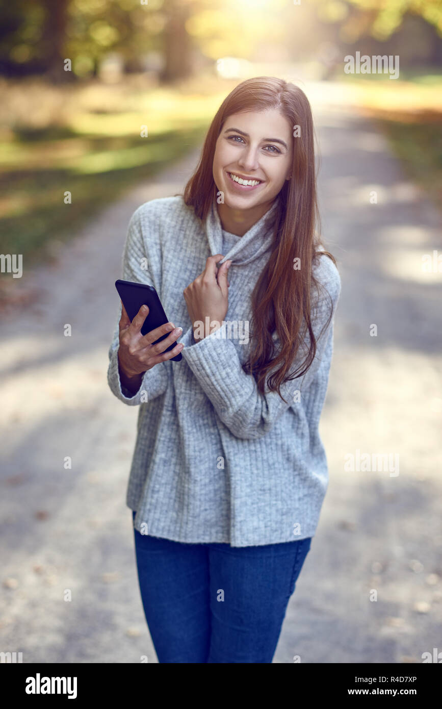 Jeune femme brune en chandail gris et bleu jeans en utilisant big smartphone dans boîtier noir, tout en descendant la route de campagne. En regardant l'écran et smi Banque D'Images