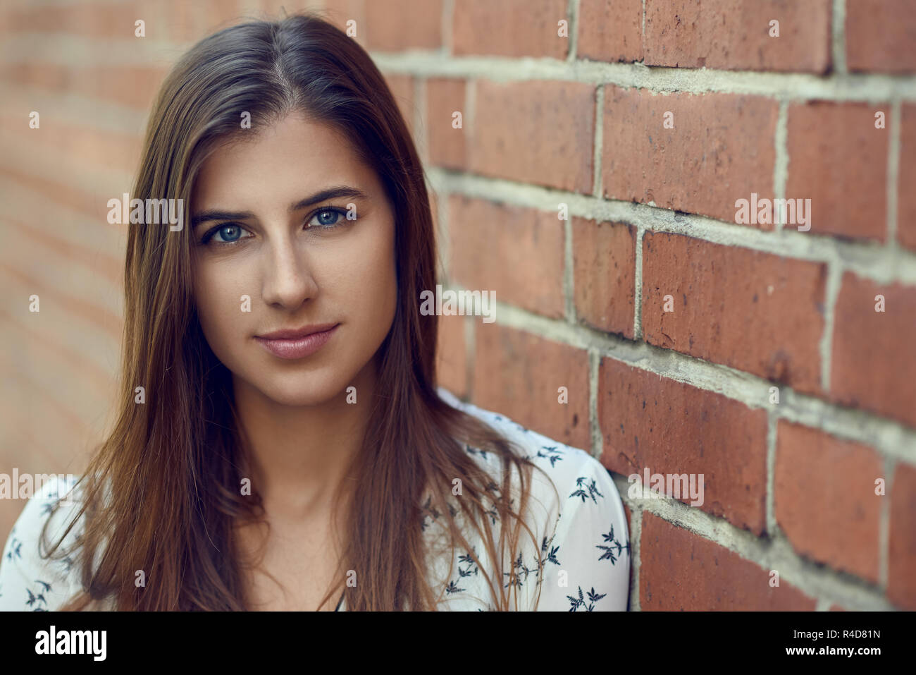 Portrait of young beautiful brunette woman with friendly joli visage, en blouse blanche, looking at camera while standing près de mur de brique rouge avec l'exemplaire Banque D'Images