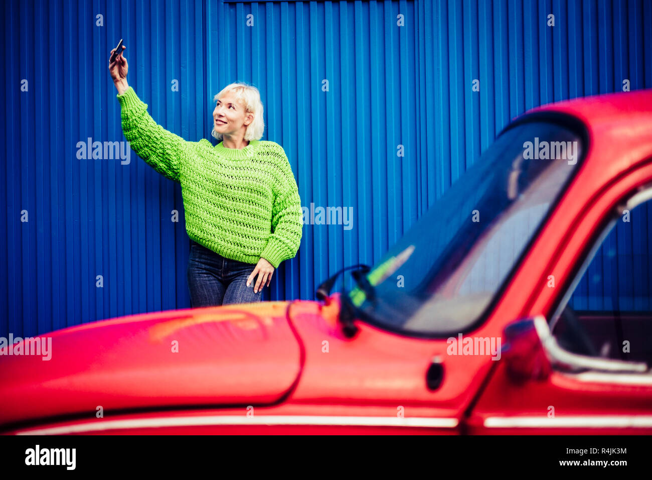 Téléphone mobile selfies happy people technologie concept avec belle blonde avec des vêtements verts et rouge et bleu voiture vintage wall en arrière-plan. M moderne Banque D'Images