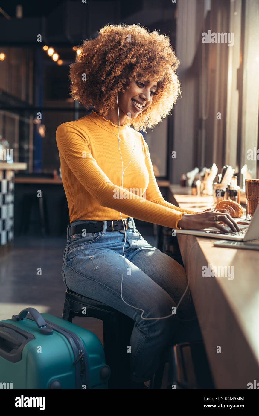 Smiling afro-américain femme qui voyage assis à un café à travailler sur ordinateur portable tout en écoutant de la musique. Femme en vacances, faire une vidéo Banque D'Images