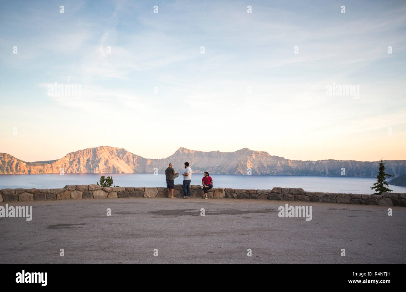 Vue éloignée sur trois jeunes gens assis sur un mur de roche à court donnant sur un grand lac de montagne au coucher du soleil, Crater Lake, Oregon, USA Banque D'Images