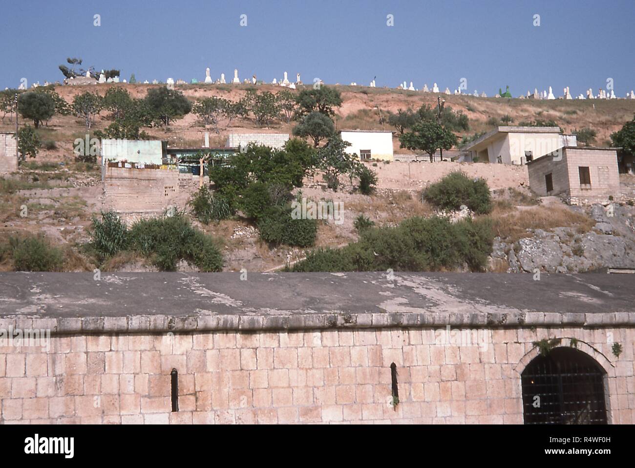 Vue sur les maisons construites à flanc de colline dans la région de Qalaat Al-Madiq, une ville habitée à côté de l'ancienne cité d'Apamée, Syrie, Juin, 1994. Au bord inférieur est le mur extérieur et la cour intérieure de l'ère ottomane entourant l'Apamée musée archéologique. () Banque D'Images