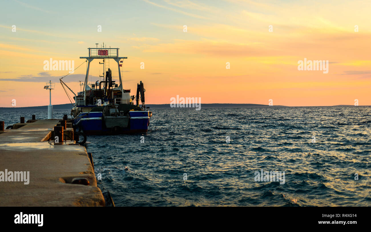 Bateau de pêche amarré ou accoste au quai au coucher du soleil. Navire de pêche commerciale dans la soirée à la tombée de la nuit avec le coucher du soleil et la couleur orange de l'île de Silba, Croatie. Banque D'Images