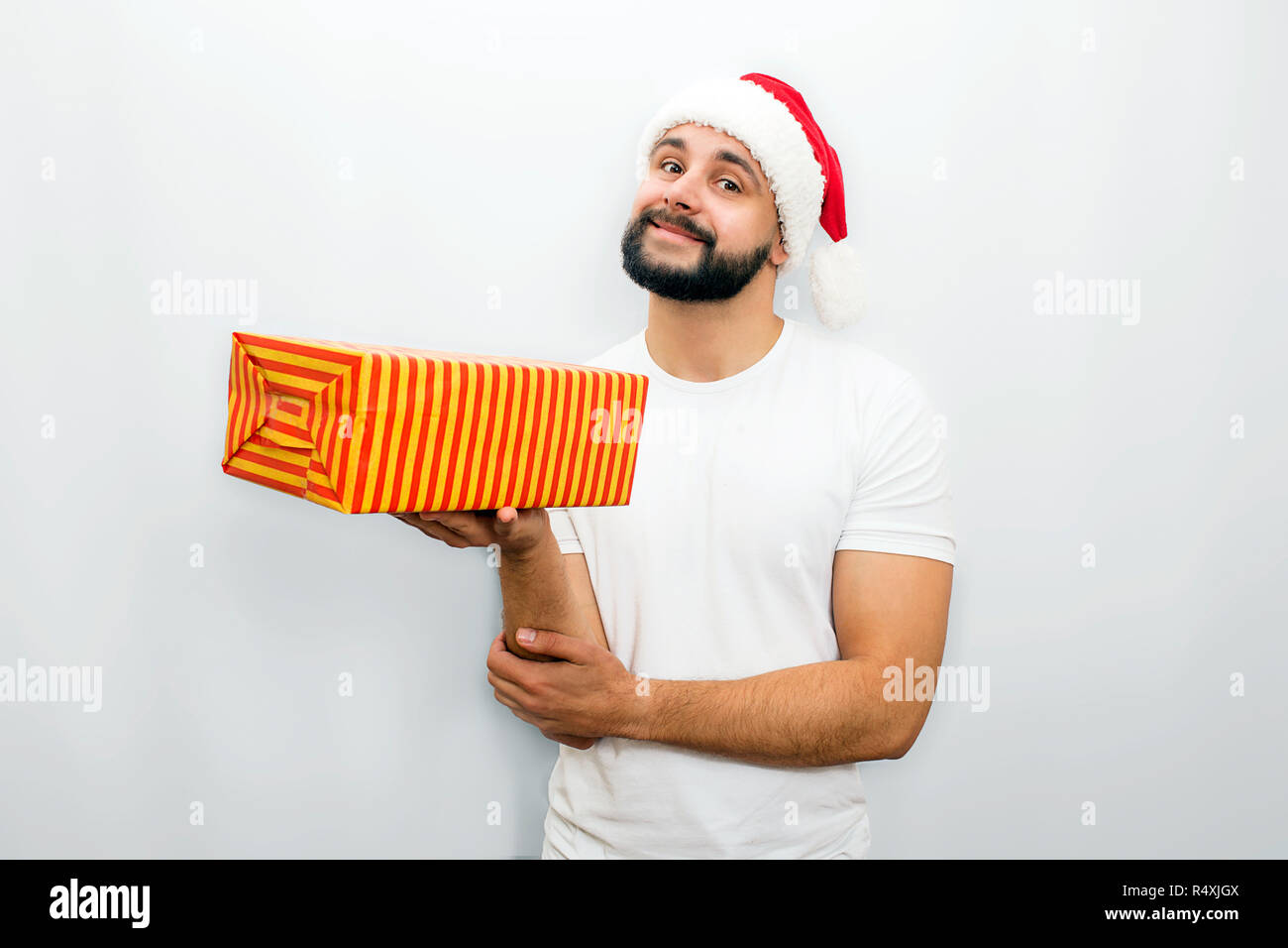 Beau barbu dans red hat se tient et est titulaire d'orange box avec présents dans une main. Il a l'air sur la caméra et sourit un peu isolé sur fond blanc. Banque D'Images