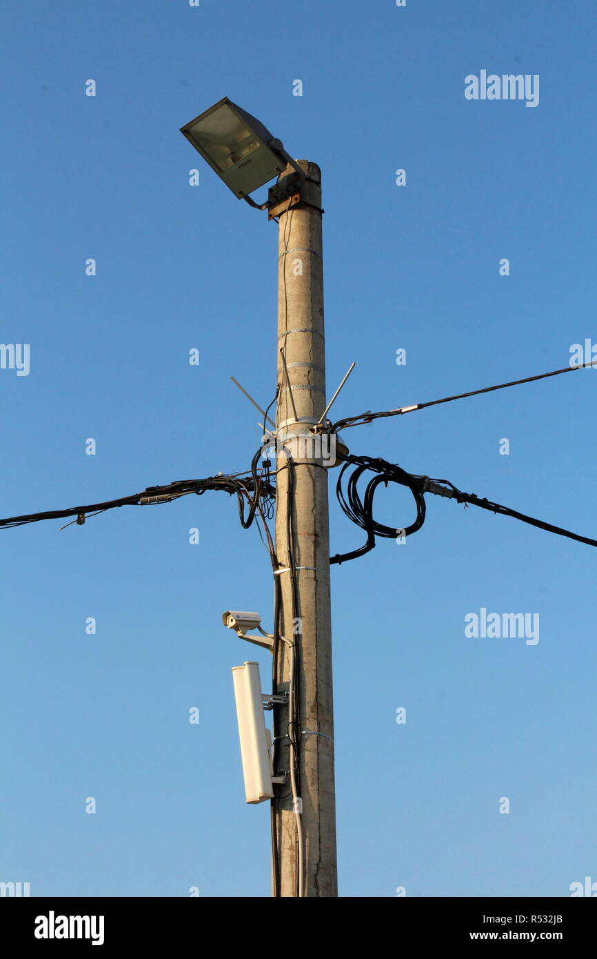 Poteau de béton avec plusieurs fils électriques noirs, petite caméra blanche, tour de téléphonie cellulaire émetteur, pointes de métal pour empêcher l'escalade Banque D'Images