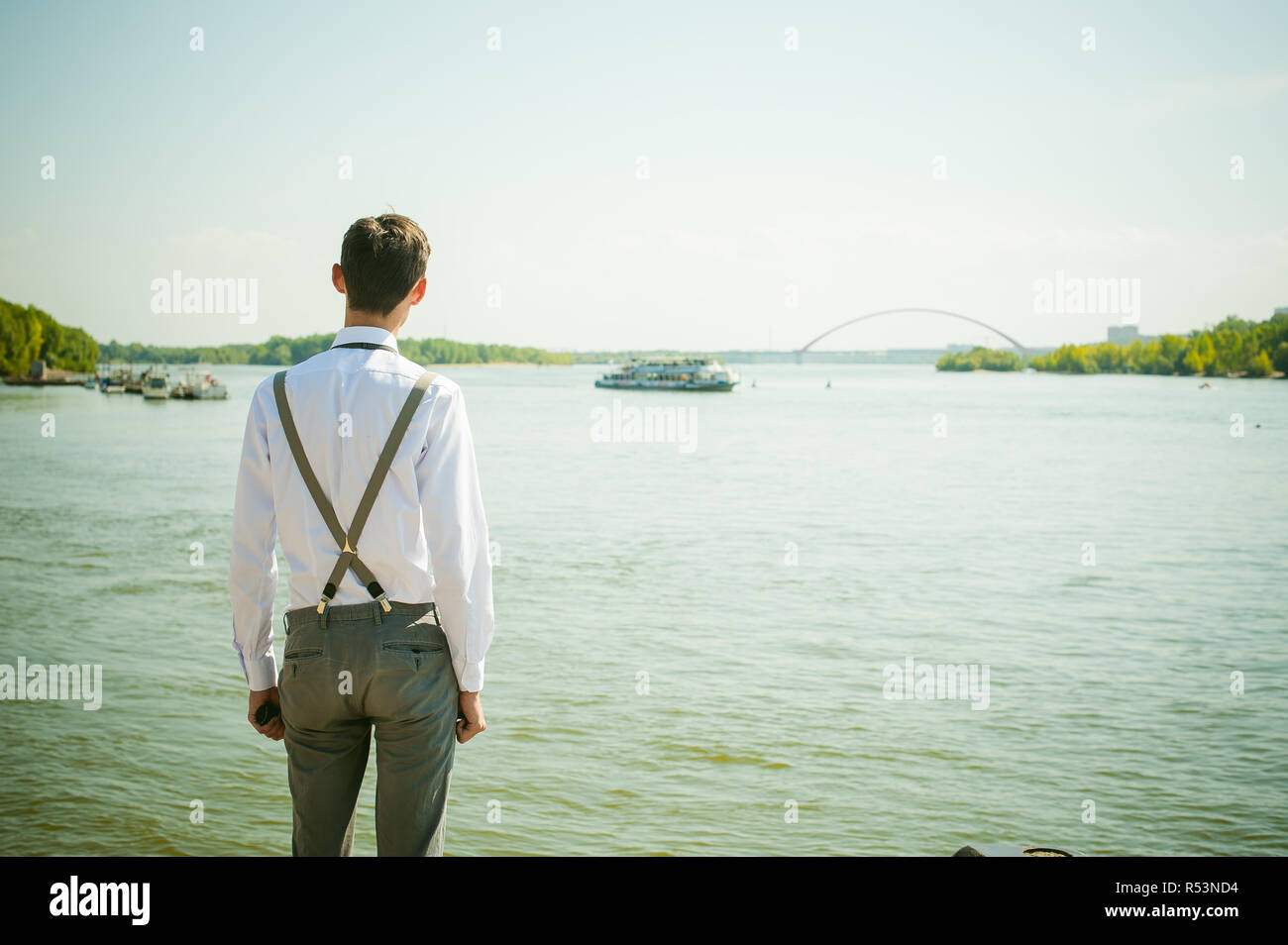 Jeune homme filiforme, élégamment habillé élégamment en chemise blanche, pantalon gris avec bretelles et dentelle. À la suite, à l'eau de la rivière et du pont de bateau Banque D'Images