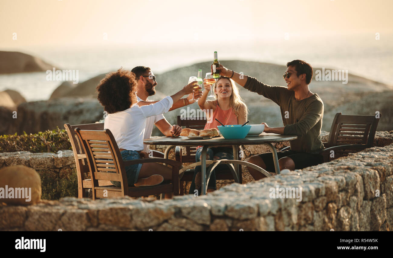 Quatre friends toasting drinks assis sur une table à manger en plein air près du littoral. En vacances amis multiethnique ayant du plaisir de boire du vin et snack Banque D'Images
