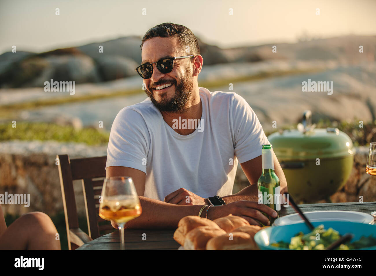 Close up of a smiling man in sunglasses assis à une table avec de la nourriture et des boissons. Homme sur une maison de vacances dans un emplacement à la campagne s'amusant avec des amis ea Banque D'Images