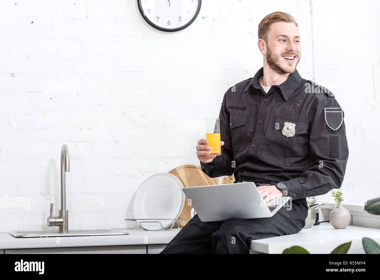 Bel homme en uniforme de policier assis sur une table de cuisine, de boire du jus d'orange et à l'aide d'ordinateur portable Banque D'Images