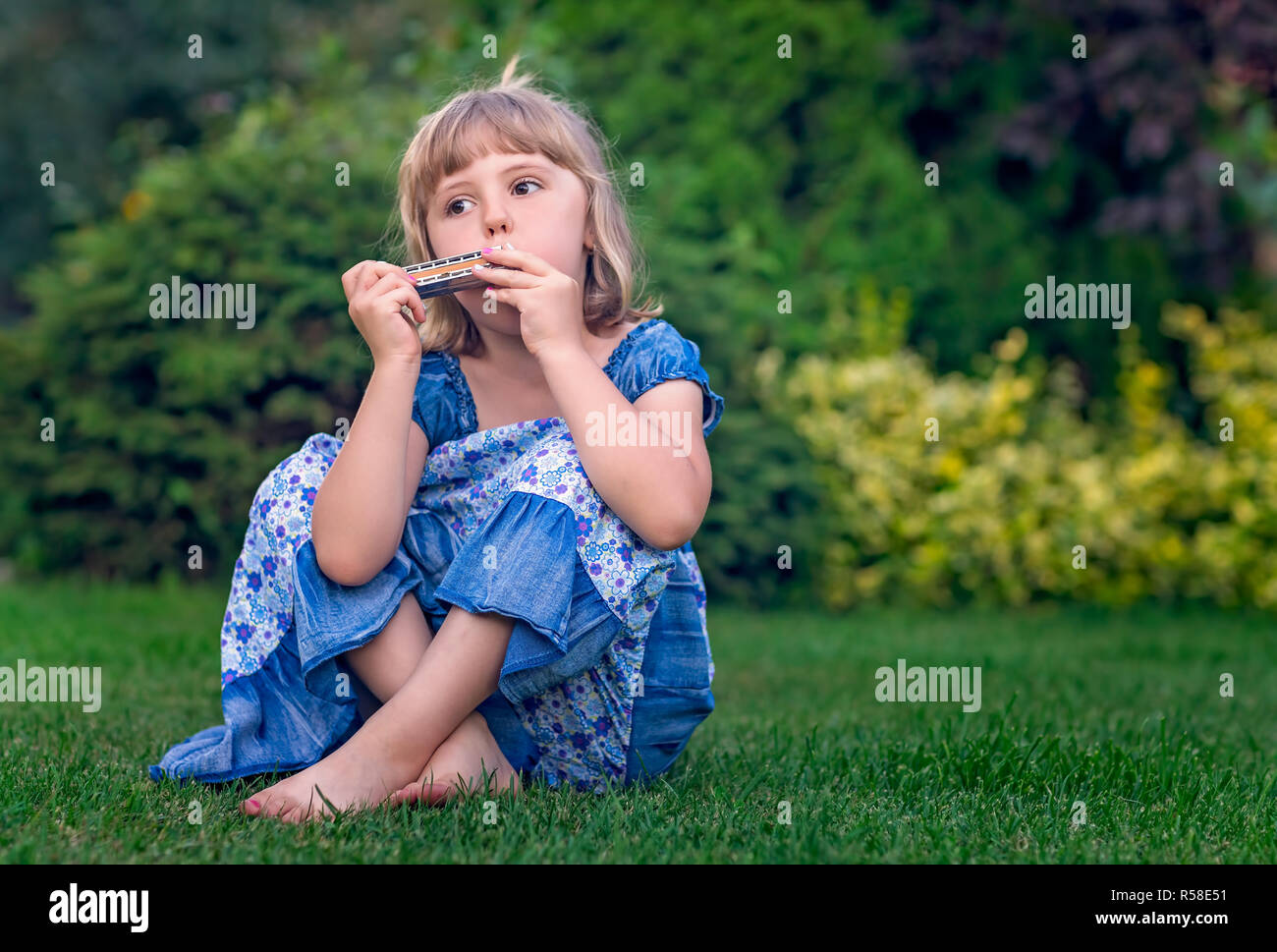 Cute girl habillé en robe bleue assise sur l'herbe dans le jardin et de s'amuser avec son petit harmonica Banque D'Images