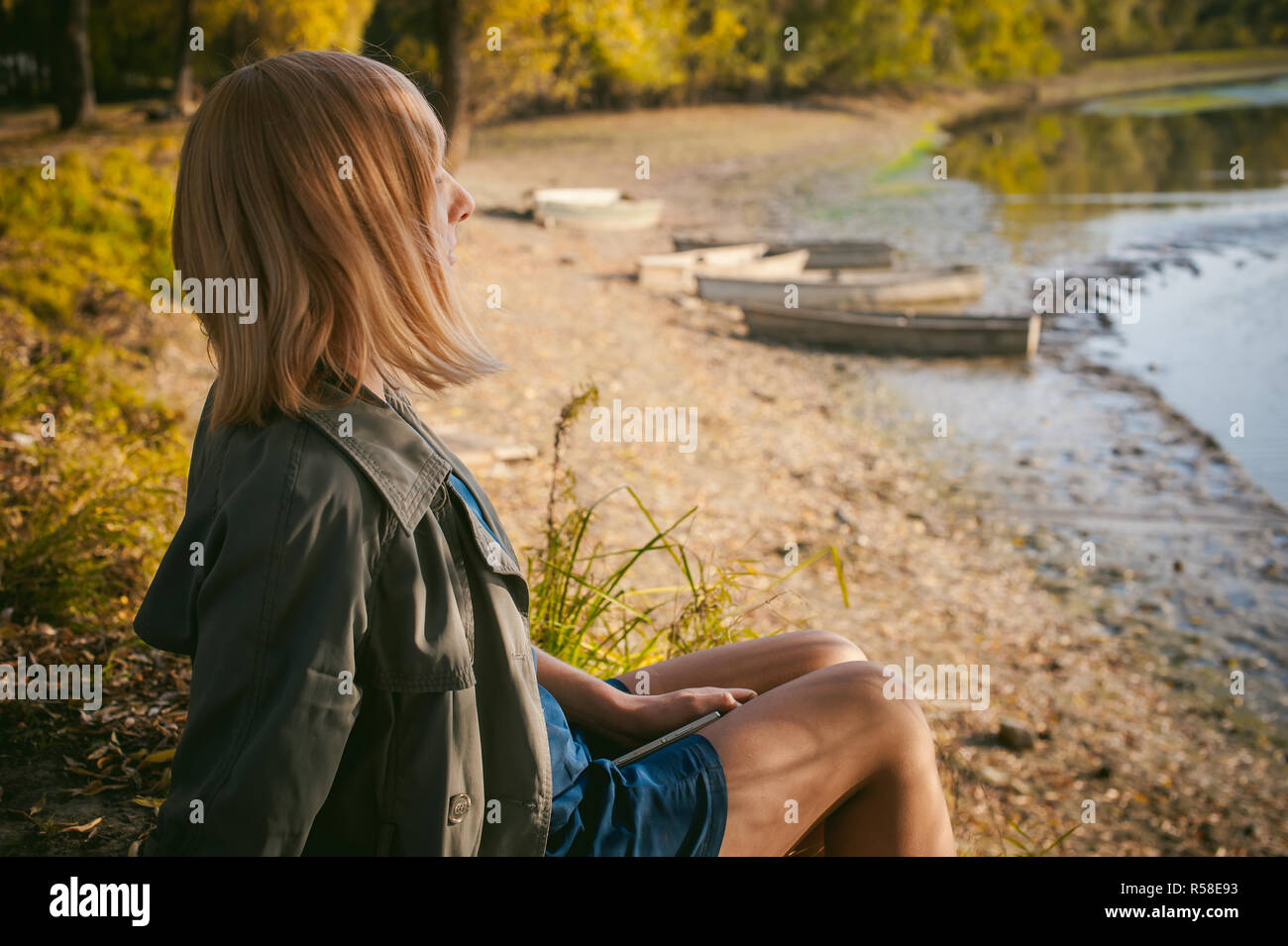 Très jolie jeune femme en sneakers, bas nylon et manteau, siège au bord de la rivière, sur des feuilles jaunes, appréciant la chaleur du soleil d'automne Banque D'Images