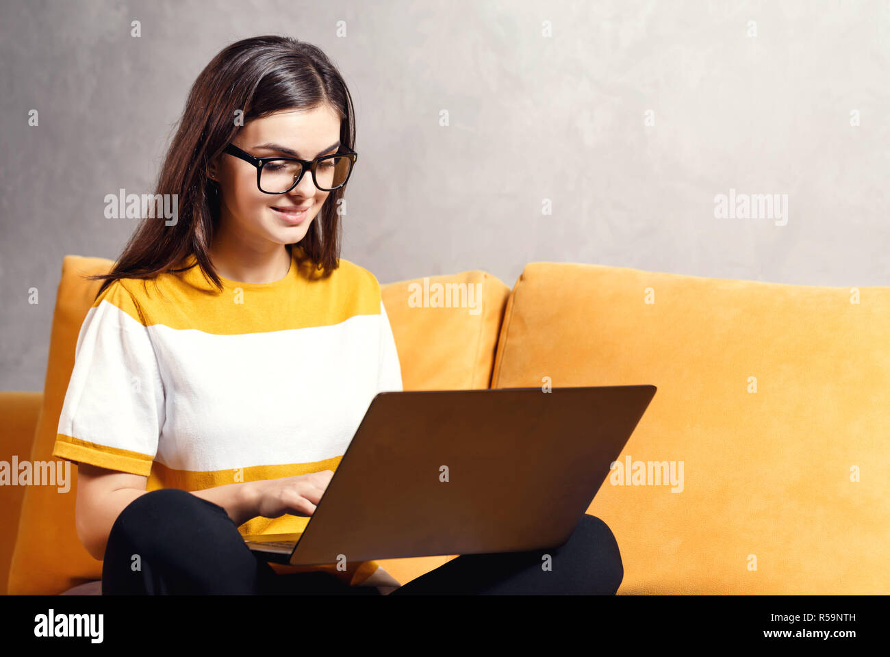 Jolie fille brune aux cheveux longs portant des lunettes assis sur le  canapé jaune à la lumière salle de séjour et de l'écriture son examen test  sur l'ordinateur portable, la technologie moderne,
