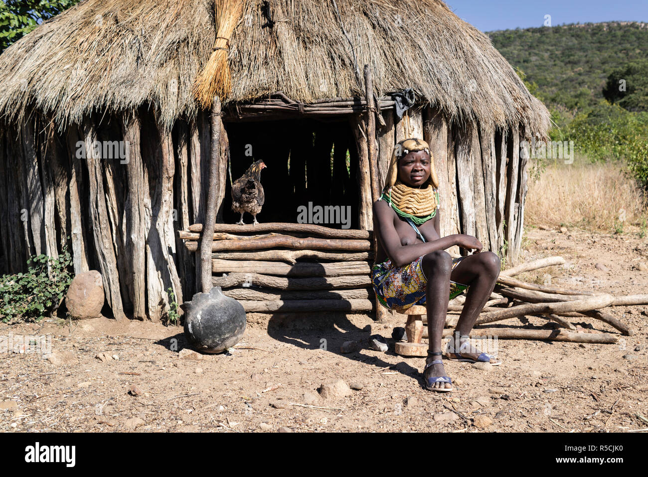 Portrait d'une jeune fille Muila assis devant sa hutte alors qu'un poulet soit saisie de l'intérieur. Banque D'Images