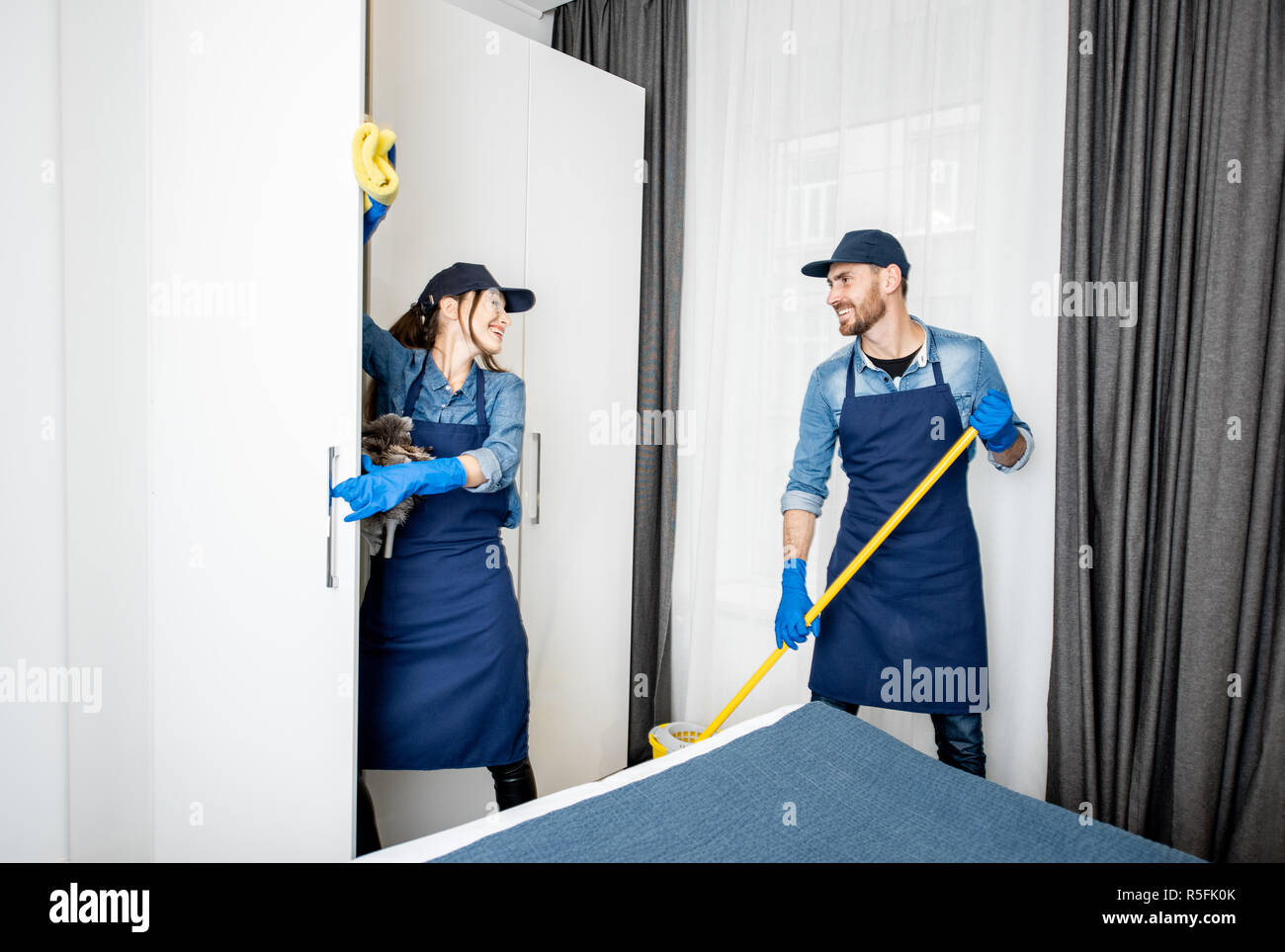 Les nettoyeurs professionnels en uniforme bleu lave-de-chaussée et d'essuyage blanc meubles de la chambre d'hôtel ou un appartement une chambre. Le concept de service de nettoyage Banque D'Images