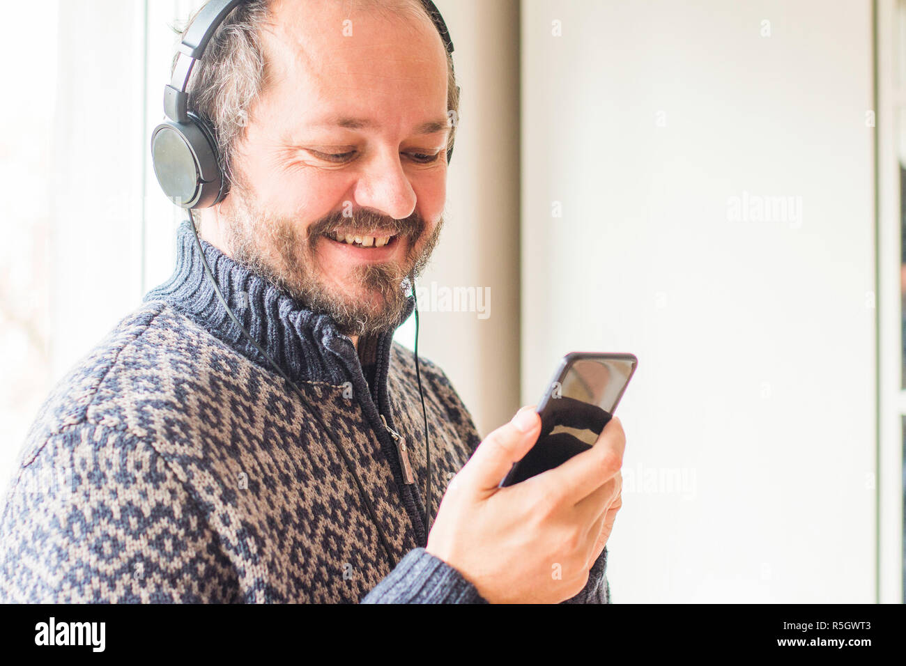 L'homme souriant l'âge moyen d'écouter de la musique avec un casque tout en vous tenant à côté de fenêtre à la maison et holding smart phone, tête et épaules Banque D'Images
