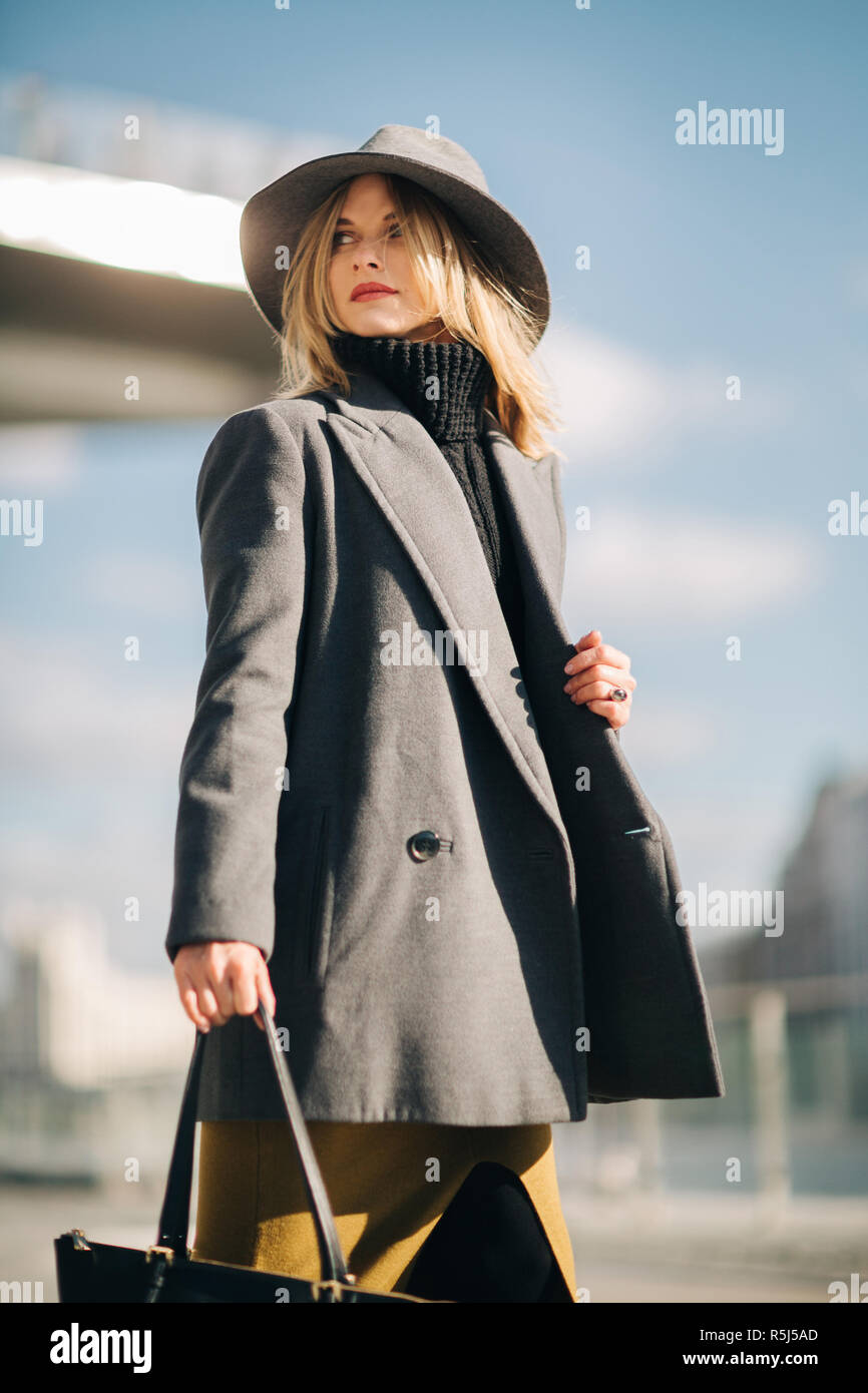 Photo ci-dessous de jeune blonde en manteau gris et un chapeau contre le ciel bleu et la construction Banque D'Images