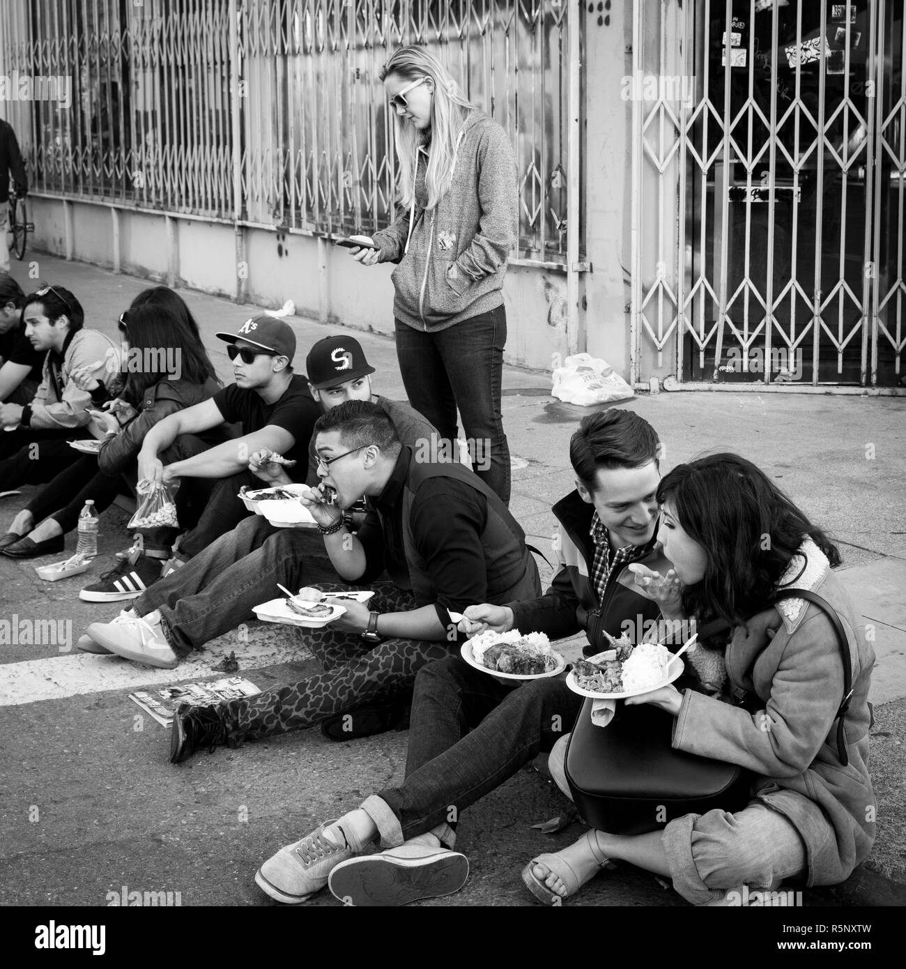 OAKLAND, CA, le 6 juin 2014 : Millénaire assis sur la bordure de la rue de manger la nourriture à un mois à l'art gallery de l'art appelé hop murmure. Carré noir et blanc. Banque D'Images