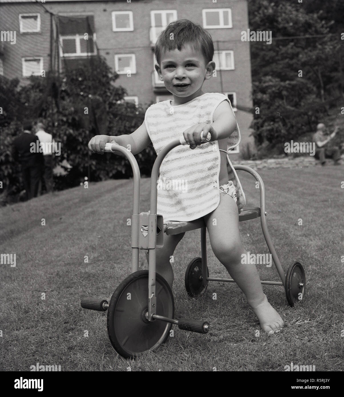 Années 1960, historique, un bébé garçon sur un tour-sur-métal produits tricycle sur une pelouse à l'extérieur d'un bloc d'appartements, England, UK. Tricycles ou trikes aider les enfants avec l'apprentissage de l'équilibre et la coordination. Banque D'Images