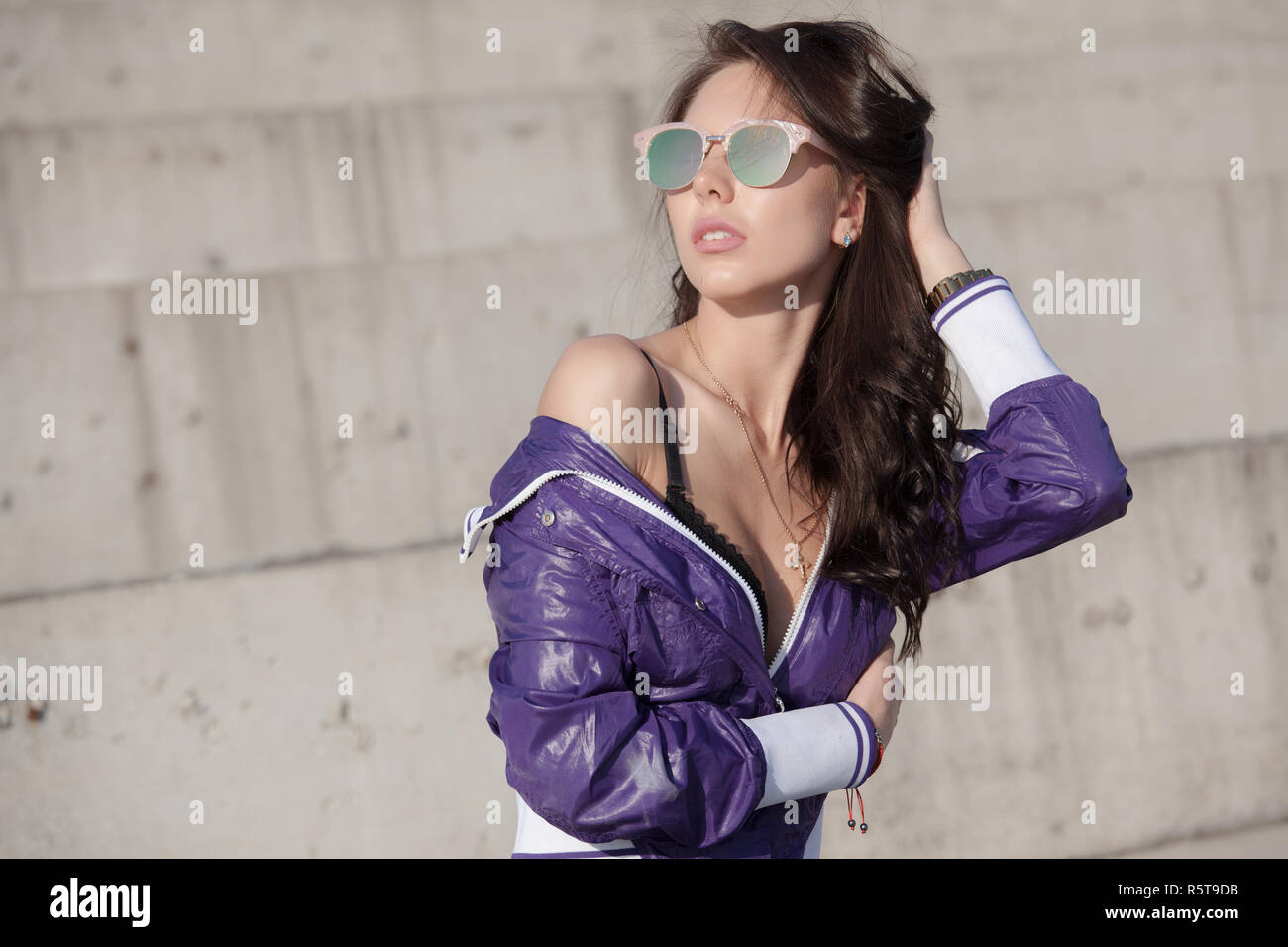 Jeune femme à la mode en lunettes de soleil, coupe-vent et un jean court  lilas. Se repose sur une structure en béton, épaule nue. La mode féminine.  Mode de vie urbain Photo