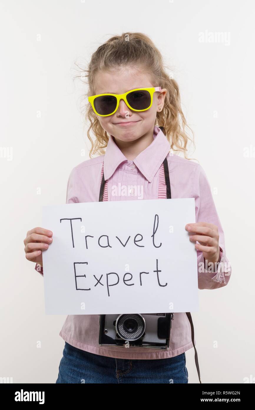 Peu d'enfants, spécialiste de la fille avec un appareil photo en lunettes holding feuille de papier blanc avec le texte d'experts de voyage, fond blanc. Banque D'Images