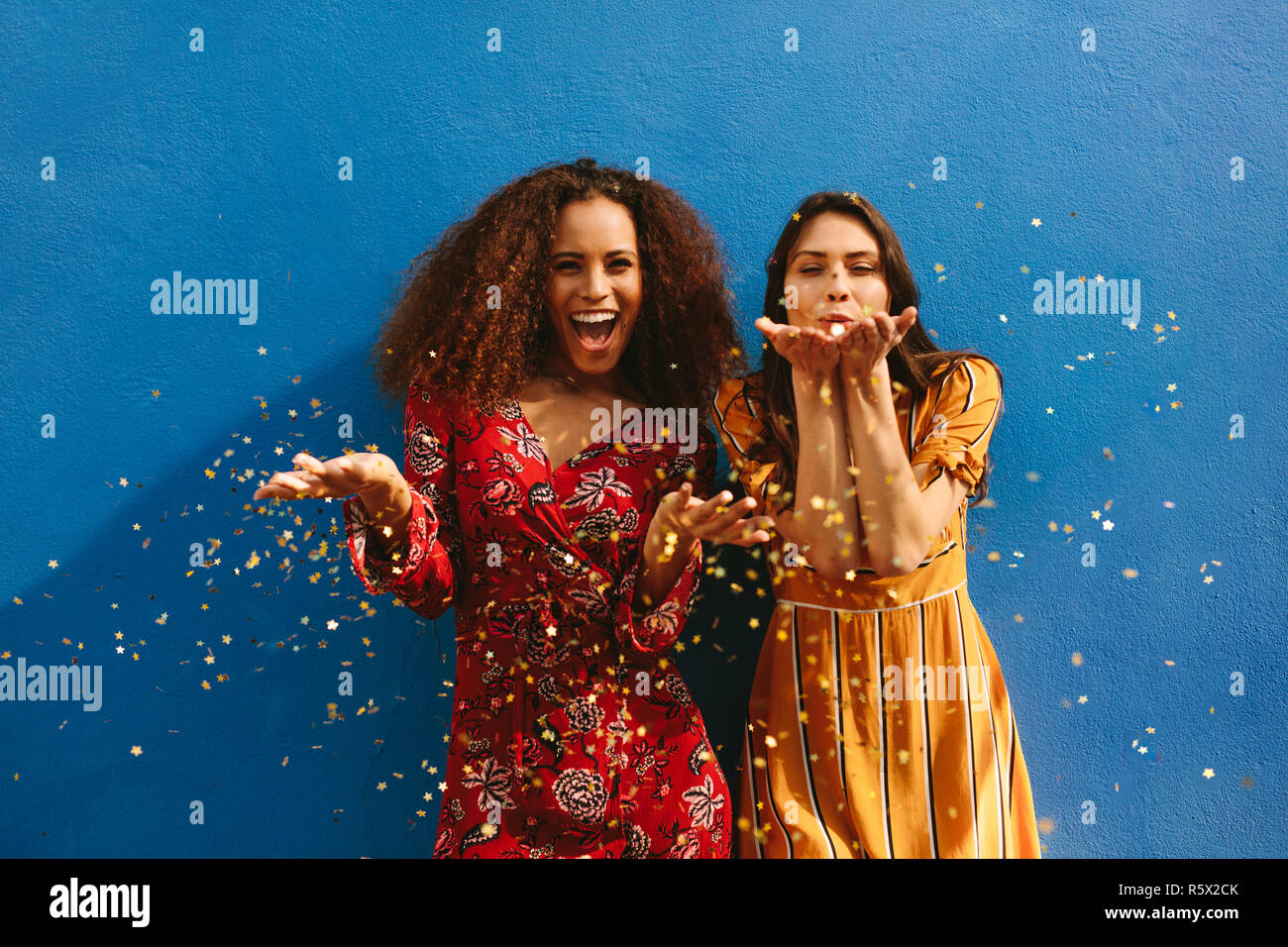 Female friends blowing off magic glitter. Les femmes excitées s'amusant contre mur bleu. Banque D'Images