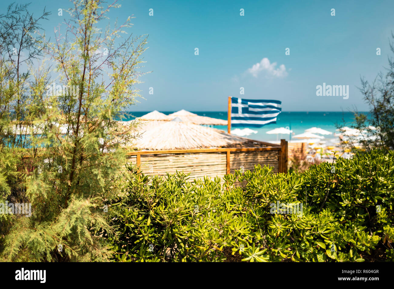 De flou artistique un drapeau grec dans le vent en face d'une plage tropicale avec parasols blancs, vu de derrière les buissons. Banque D'Images