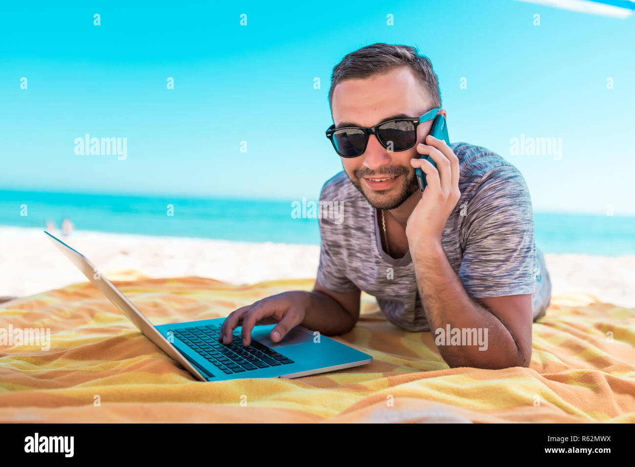 Jeune homme séduisant en lunettes de soleil avec ordinateur portable et  téléphone mobile sur plage Photo Stock - Alamy