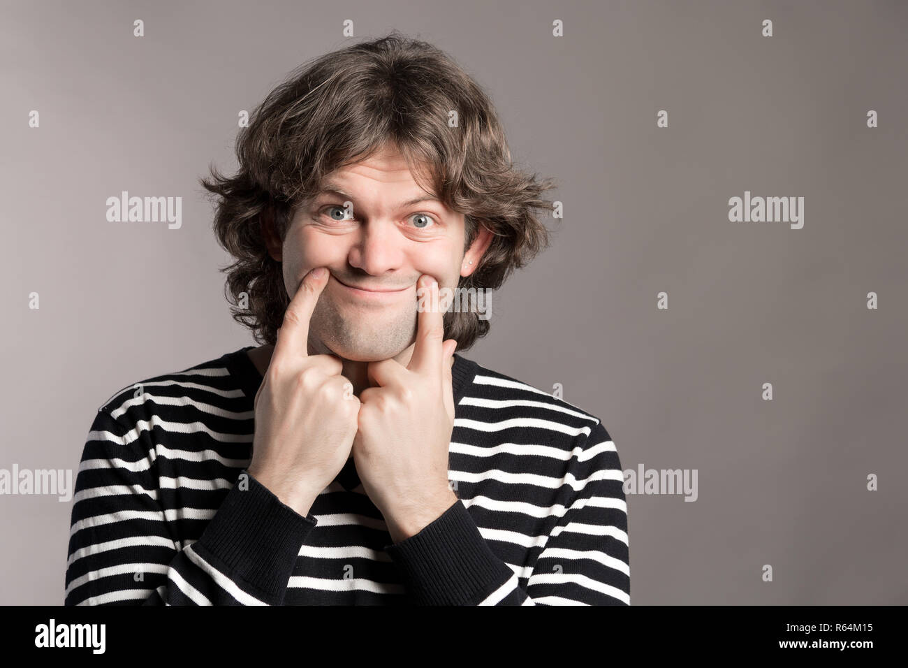 Jeune homme fait un drôle de sourire en aidant son index. Expression figure sur un fond gris de l'espace de copie. Triste dark-haired young man dressed c Banque D'Images