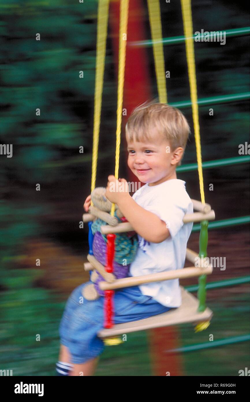 Portrait, Halbfigur, extérieur, 3-jaehriger bekleidet Junge mit weissem T-Shirt neues mit seinem Kuscheltier schaukelt flexible in einer mit Kinderschaukel Sprossen im Garten Banque D'Images
