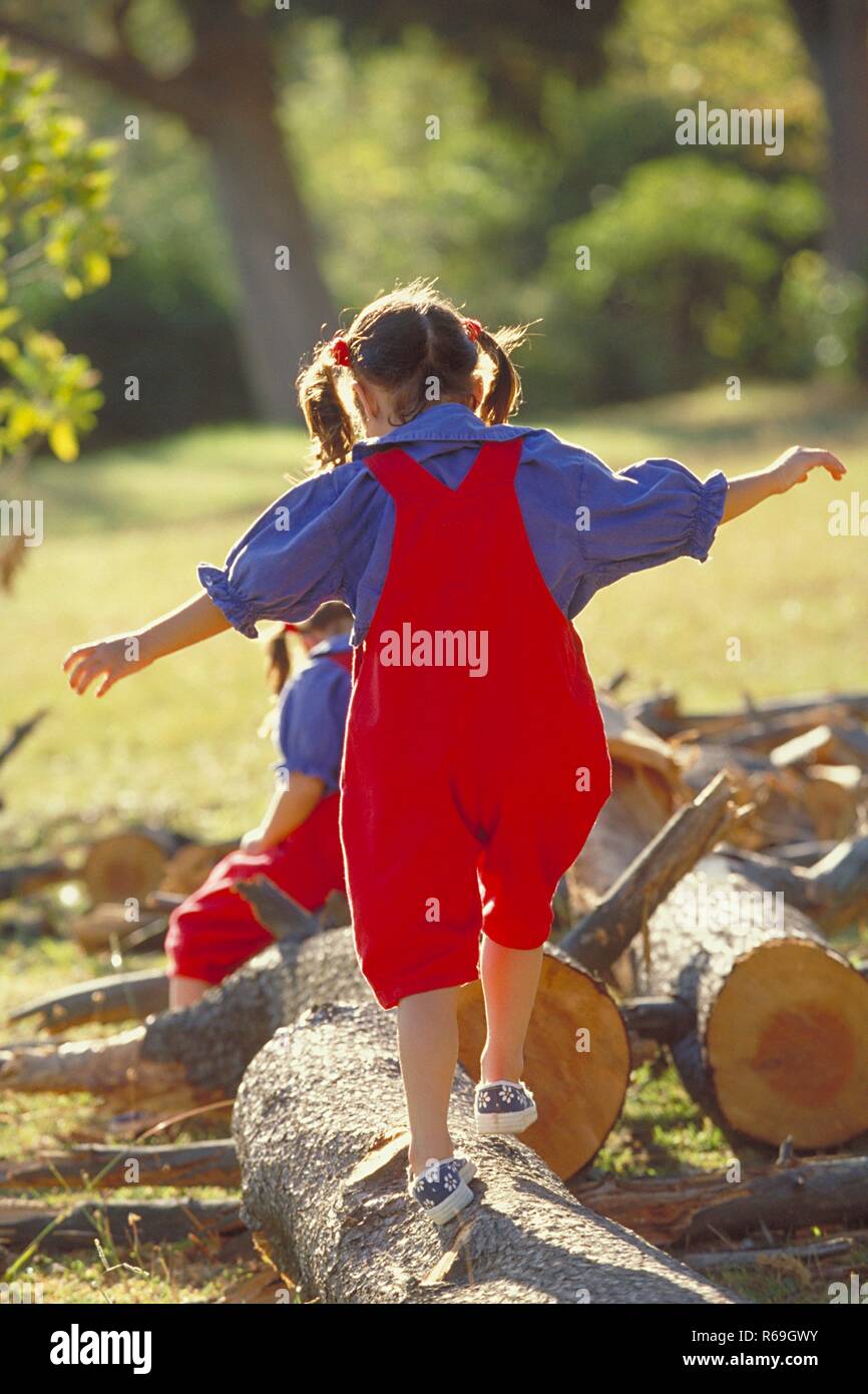 Parkszene Ganzfigur bruenette,, 2 Zwillings-Maedchen, 6 Jahre alt, bekleidet mit roter Latzhose Blouse, blauer und Turnschuhen Baumstaemme dicke ueber balancieren Banque D'Images