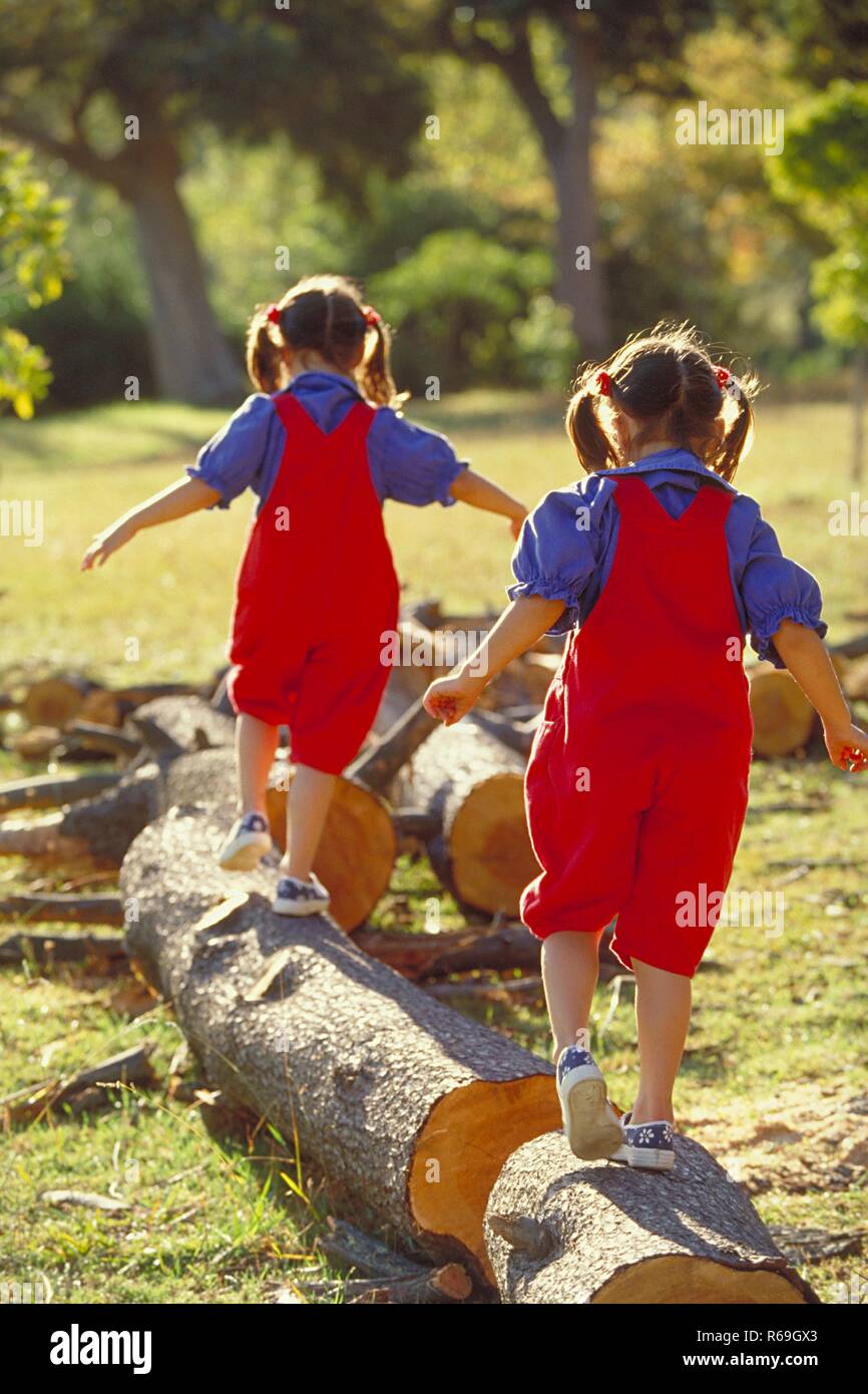 Parkszene Ganzfigur, von hinten, 2 Zwillings-Maedchen bruenette, 6 Jahre alt, bekleidet mit roter Latzhose, blauer und Blouse Turnschuhen Baumstaemme dicke ueber balancieren Banque D'Images
