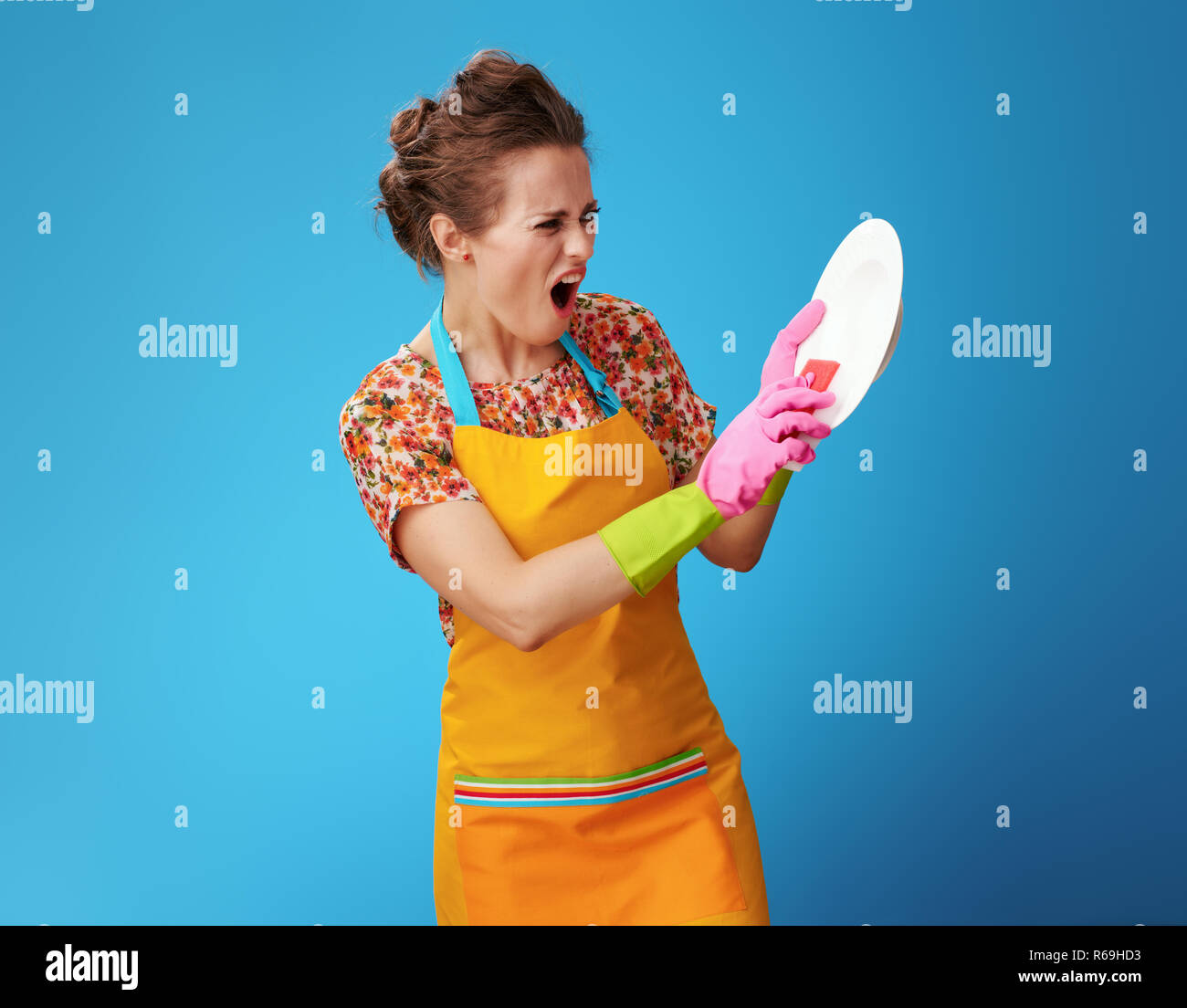 Jeune femme en colère en tablier orange avec éponge lave-vaisselle isolé sur fond bleu. Ce qui va aider à laver la vaisselle sale super ? Conseils, des détergents ou Banque D'Images