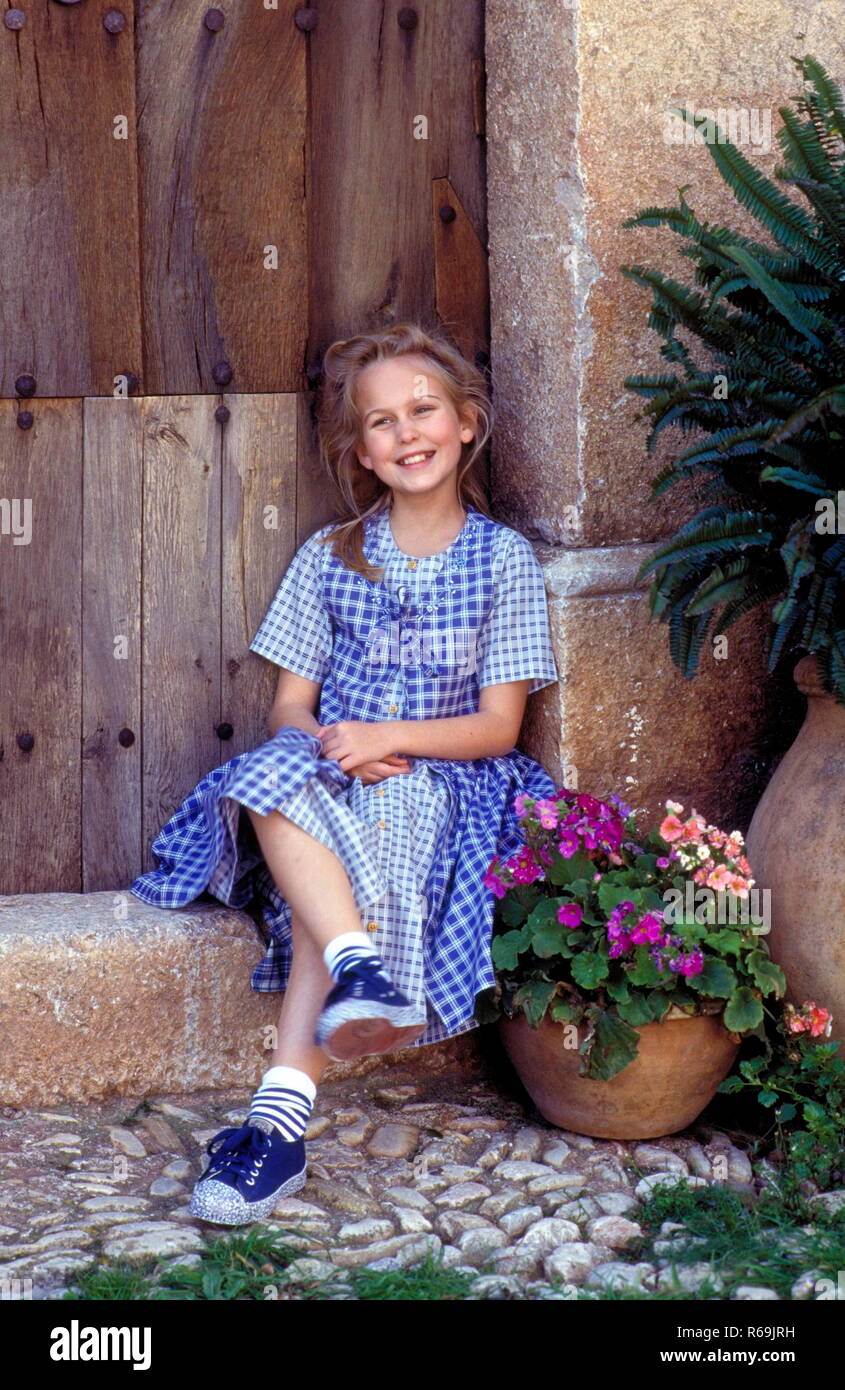 Portrait, blondes Maedchen, 10 Jahre, bekleidet-blau mit kariertem weiss  und Kleid blauen Turnschuhen lachend sitzt auf auf einer Steinstufe  Holztuer vor einer alten Photo Stock - Alamy