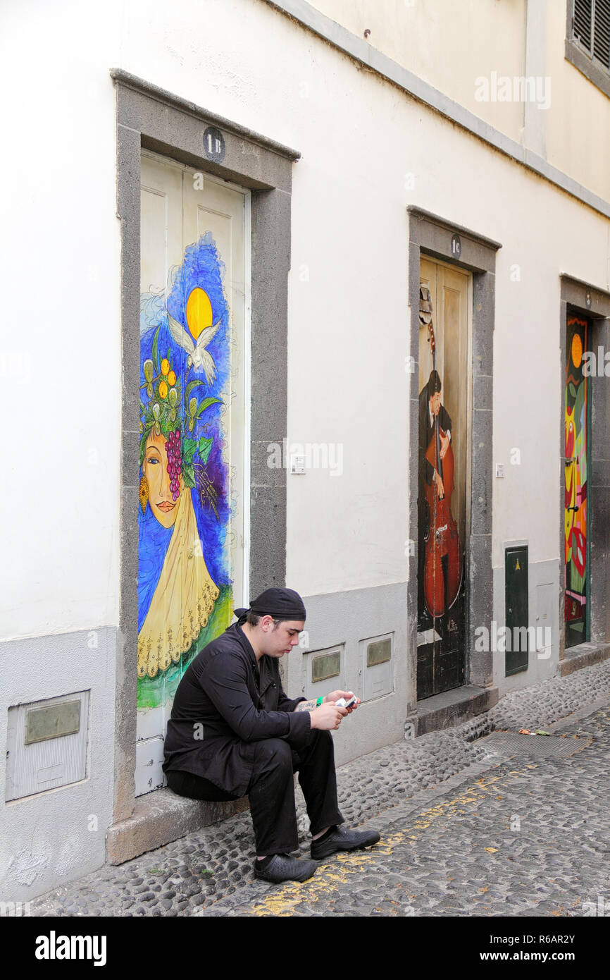 Jeune homme faire une pause en face de portes peintes dans la vieille ville de Funchal et jeter un oeil sur son smartphone Banque D'Images