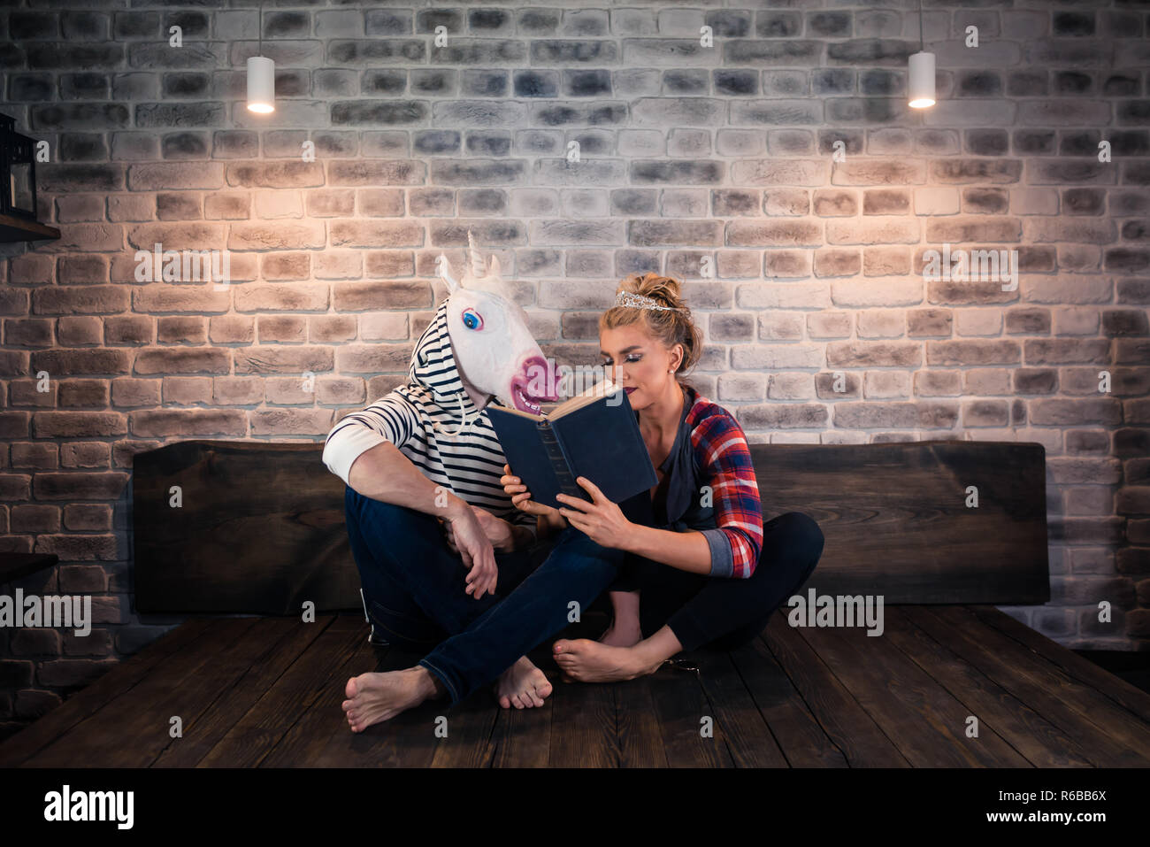 Couple rare de lire un livre dans la belle chambre. Belle jeune fille est assise sur le lit avec drôle de petit ami en masque comique. Jeune femme à l'homme bizarre. Banque D'Images