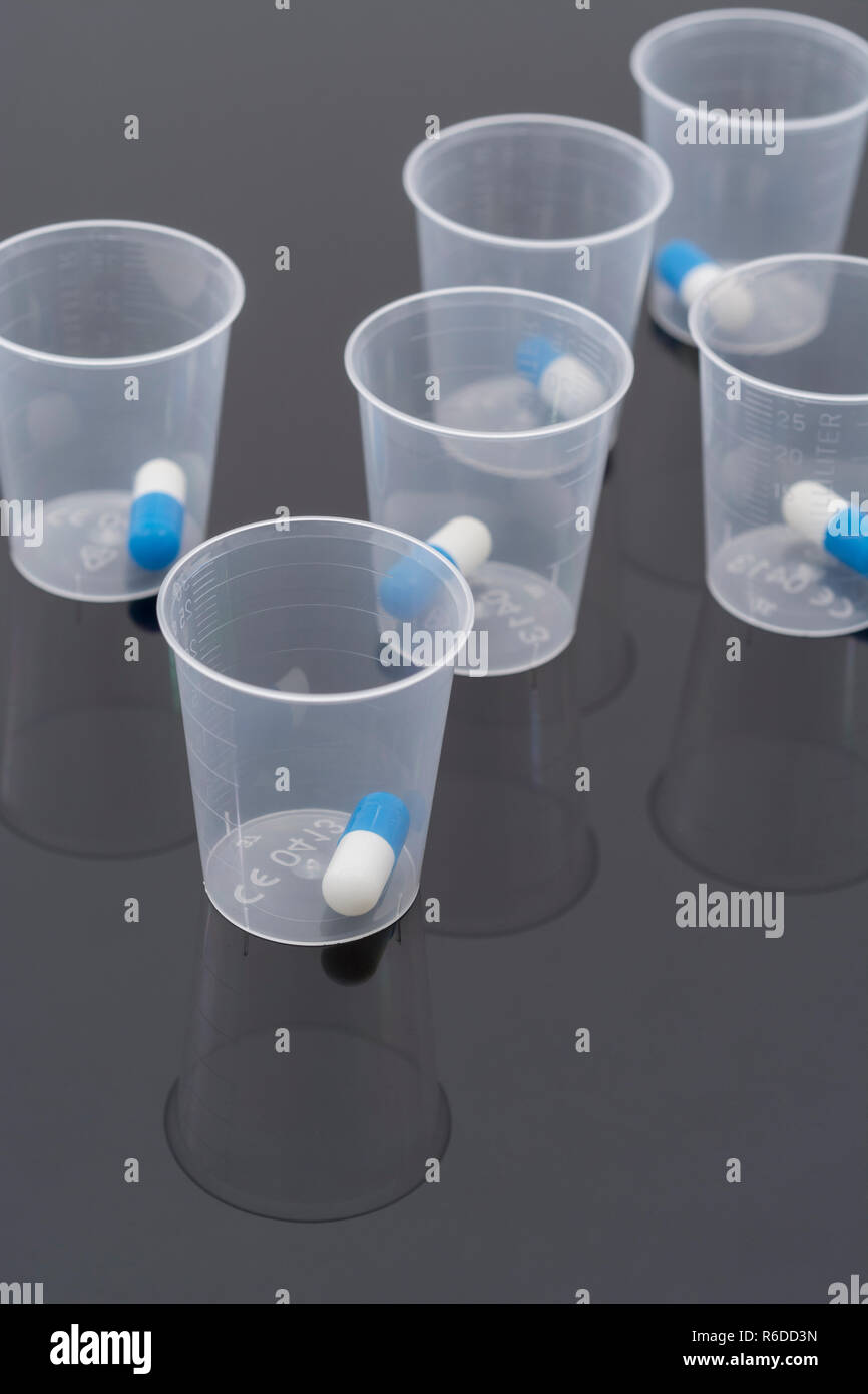 Big Pharma concept de médicaments. Capsules comprimés bleu et blanc dans une tasse à mesurer en plastique / coupe de distribution sur une surface réfléchissante. NHS possible métaphore. Banque D'Images