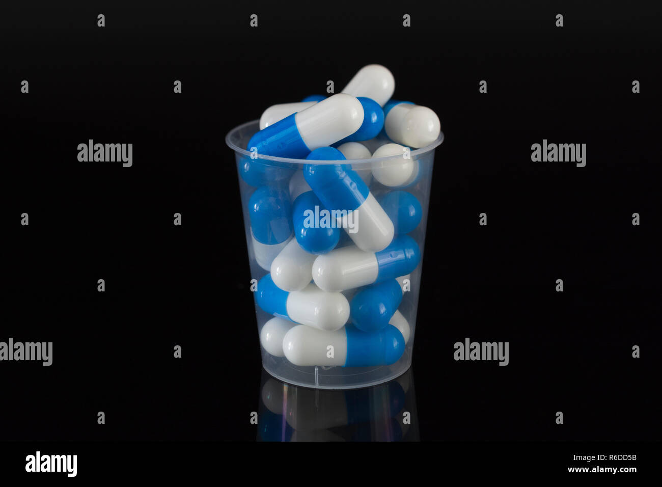 Big Pharma concept de médicaments. Capsules comprimés bleu et blanc dans une tasse à mesurer en plastique / coupe de distribution sur une surface réfléchissante. NHS possible métaphore. Banque D'Images