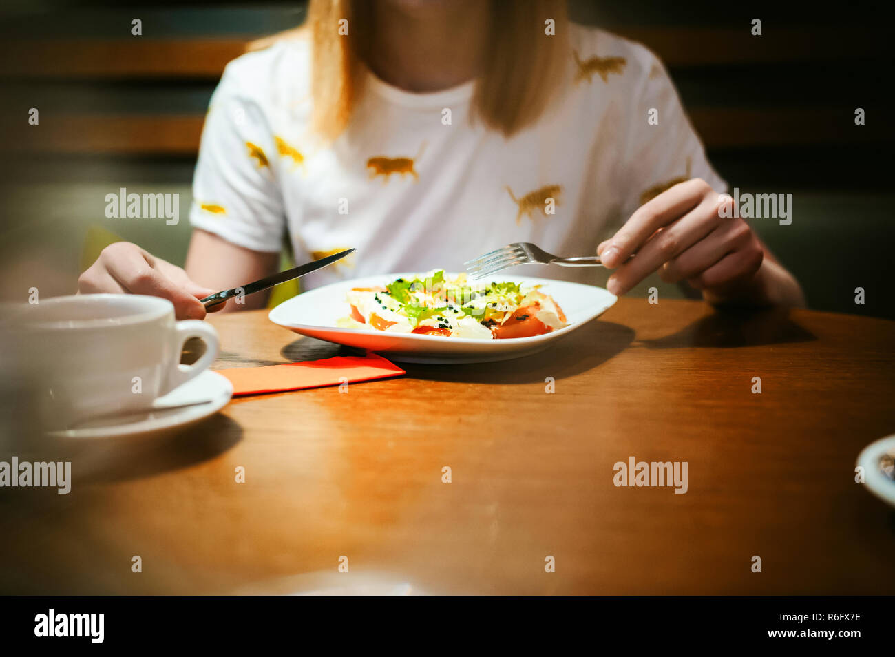 Young blonde woman wearing white T-shirt avec print, fille mange de salade à l'heure du déjeuner, assis à une table dans un café, la texture du bois stylisé d'arrière-plan éclairé avec une lumière chaude Banque D'Images