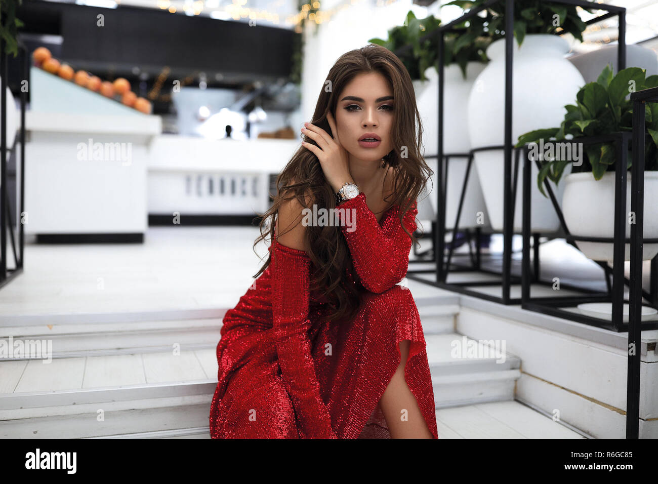 Lumineux efficace dame en robe longue rouge, assis sur des marches blanches le restaurant moderne. Maquillage sur visage, cheveux bouclés élégant situé sur les épaules. Banque D'Images