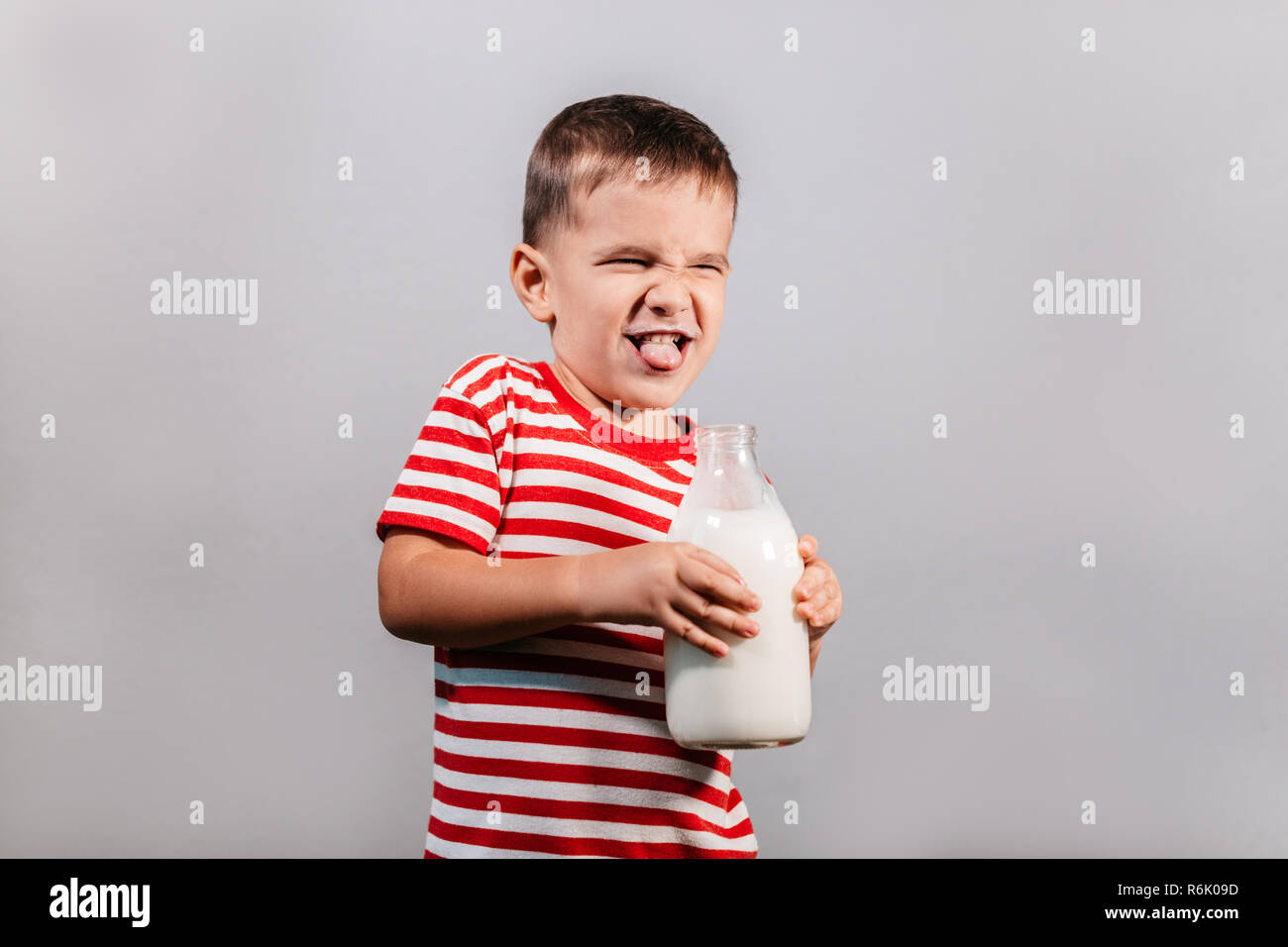 Enfant avec bouteille de lait contre fond gris. Portrait de jeune garçon avec une moustache de lait making faces isolated over grey background - studio shot. Banque D'Images