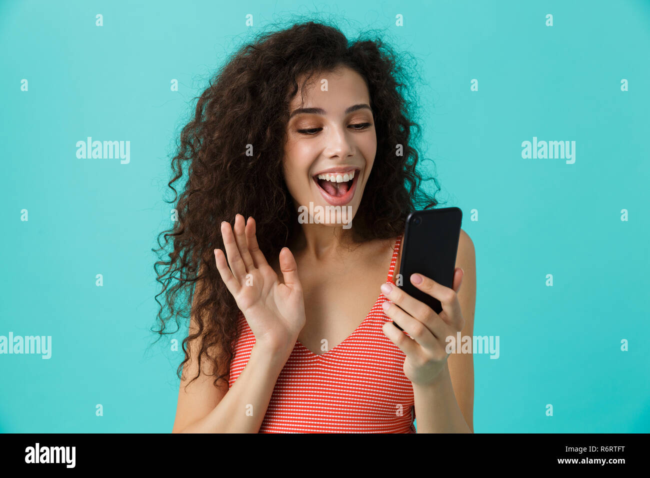 Photo de femme joyeuse 20s avec des cheveux bouclés smiling and looking at mobile phone isolé sur fond bleu Banque D'Images