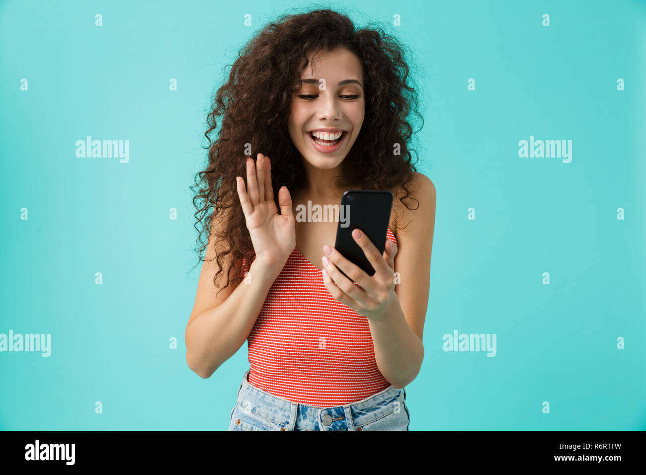 Photo de femme brune 20s avec des cheveux bouclés smiling and looking at mobile phone isolé sur fond bleu Banque D'Images