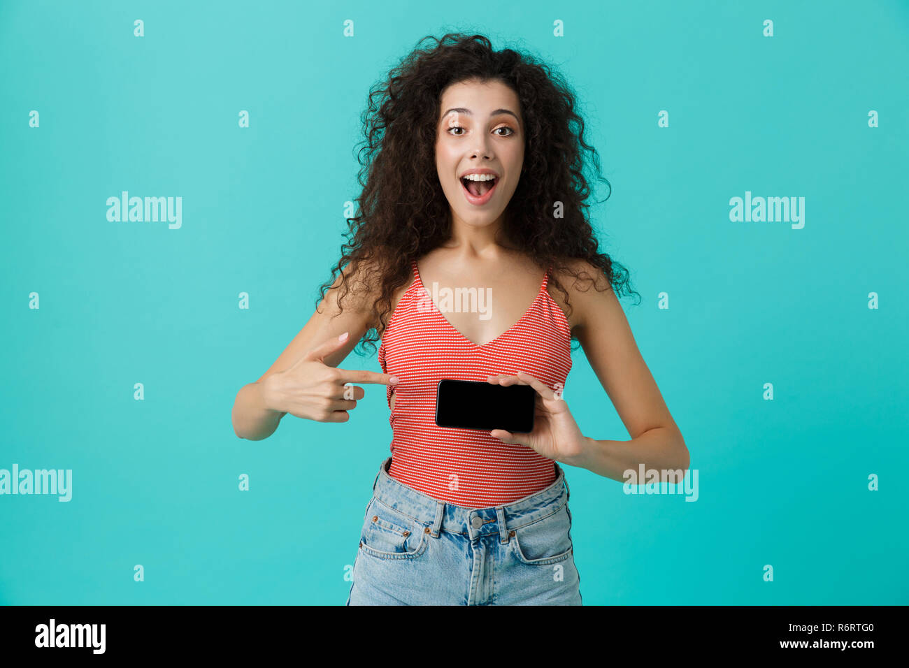 Photo de charmante femme 20s avec des cheveux bouclés souriant et holding smartphone isolé sur fond bleu Banque D'Images